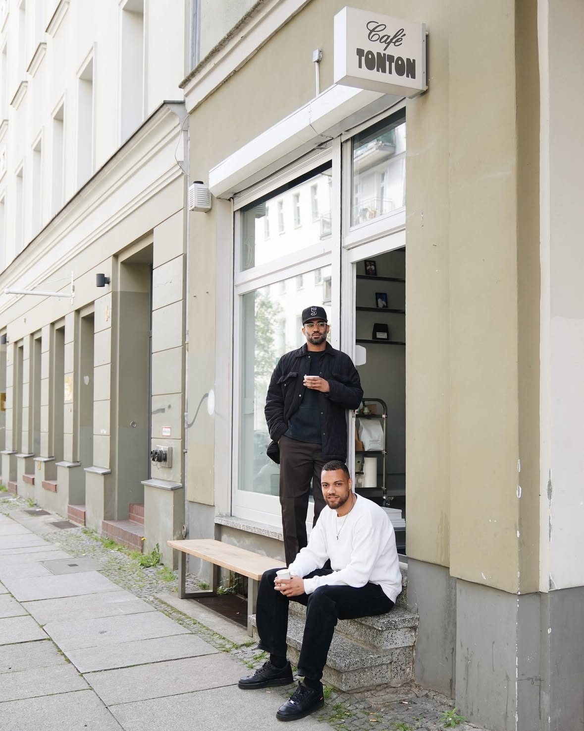 Two men outside the shopfront at Tieckstraße 1 under the hanging Café TONTON sign, holding coffee cups