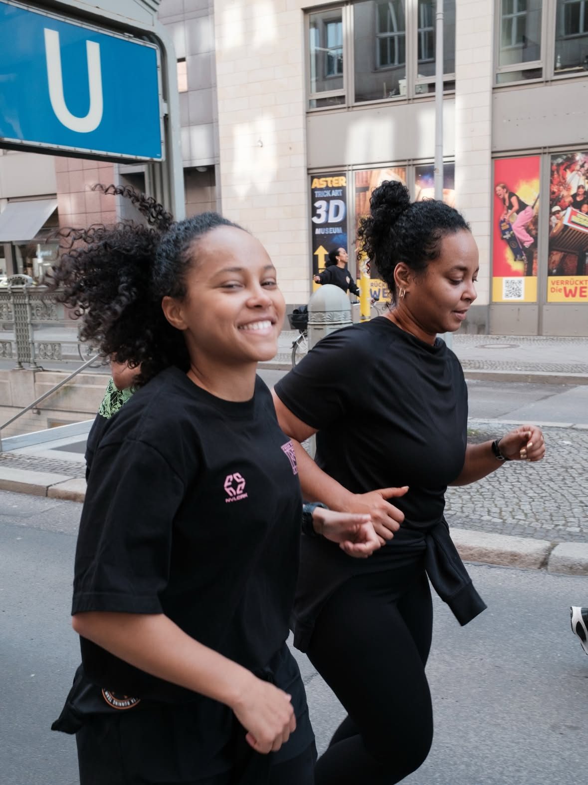 Two runners in black on a city street, passing a blue U-Bahn sign, at the Black History Month Community Run 2026.